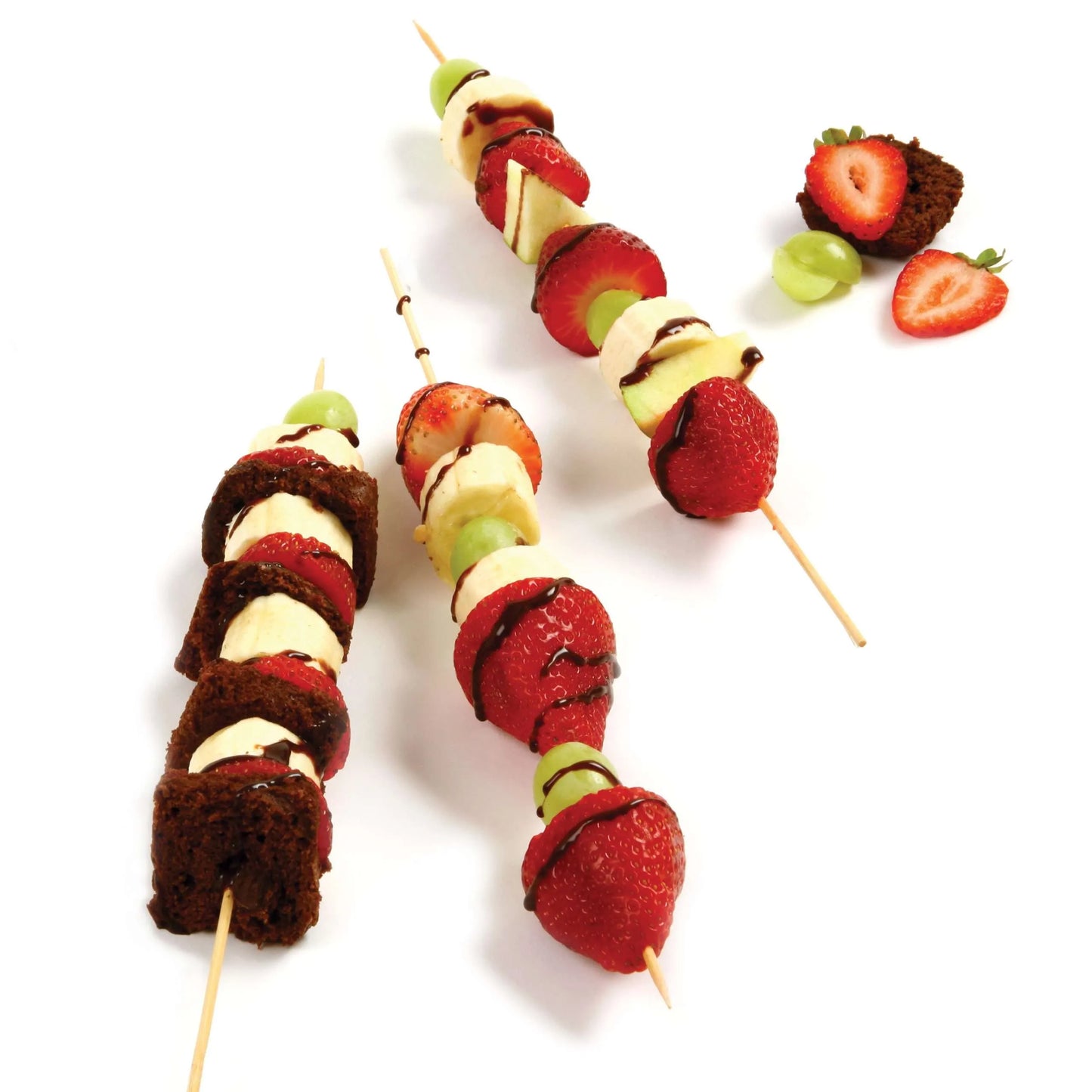 Three bamboo skewers with fresh fruit and cake bites drizzled with chocolate on a white surface.