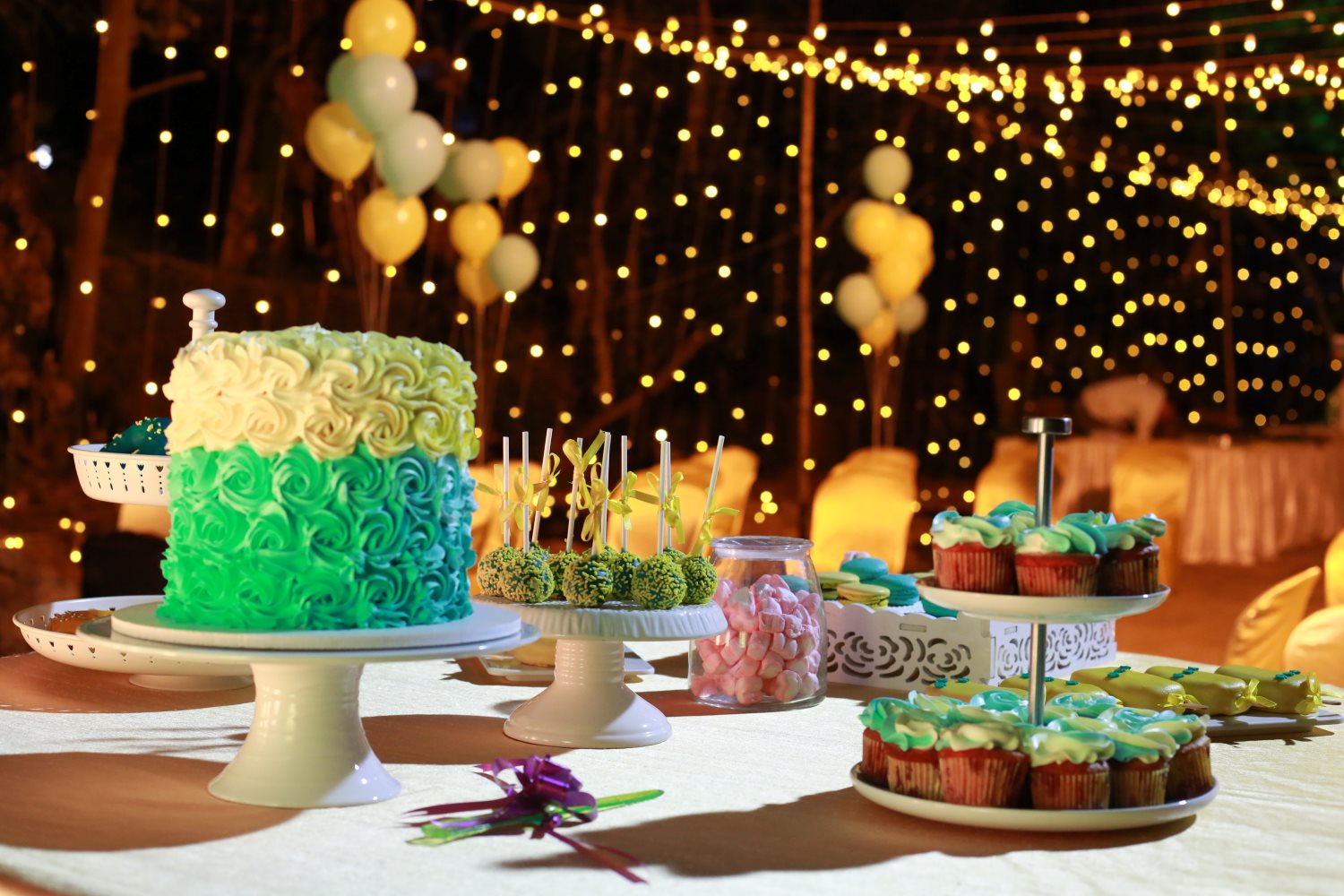 A display of a green and yellow ombre cake, cupcakes with green and blue frosting, and various other desserts at an outdoor party with string lights and balloons in the background.
