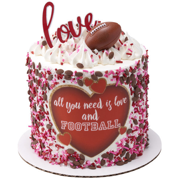 White frosted cake with pink and brown sprinkles, heart-shaped "All you need is love and football" topper, and a 3D football ring.