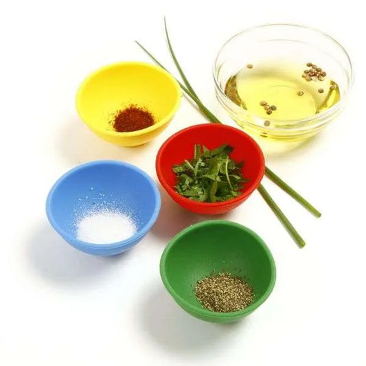 Five colorful silicone prep bowls in red, yellow, green, and blue, holding various seasonings and herbs, arranged with other fresh ingredients.