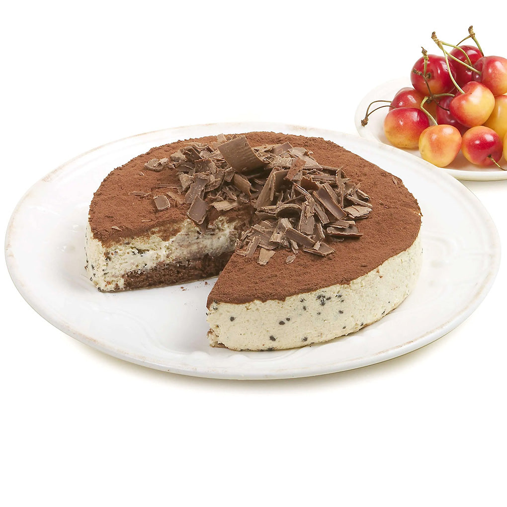 Round chocolate-topped cheesecake with a slice removed, chocolate shavings piled in the center, and red-yellow cherries displayed beside the plate.