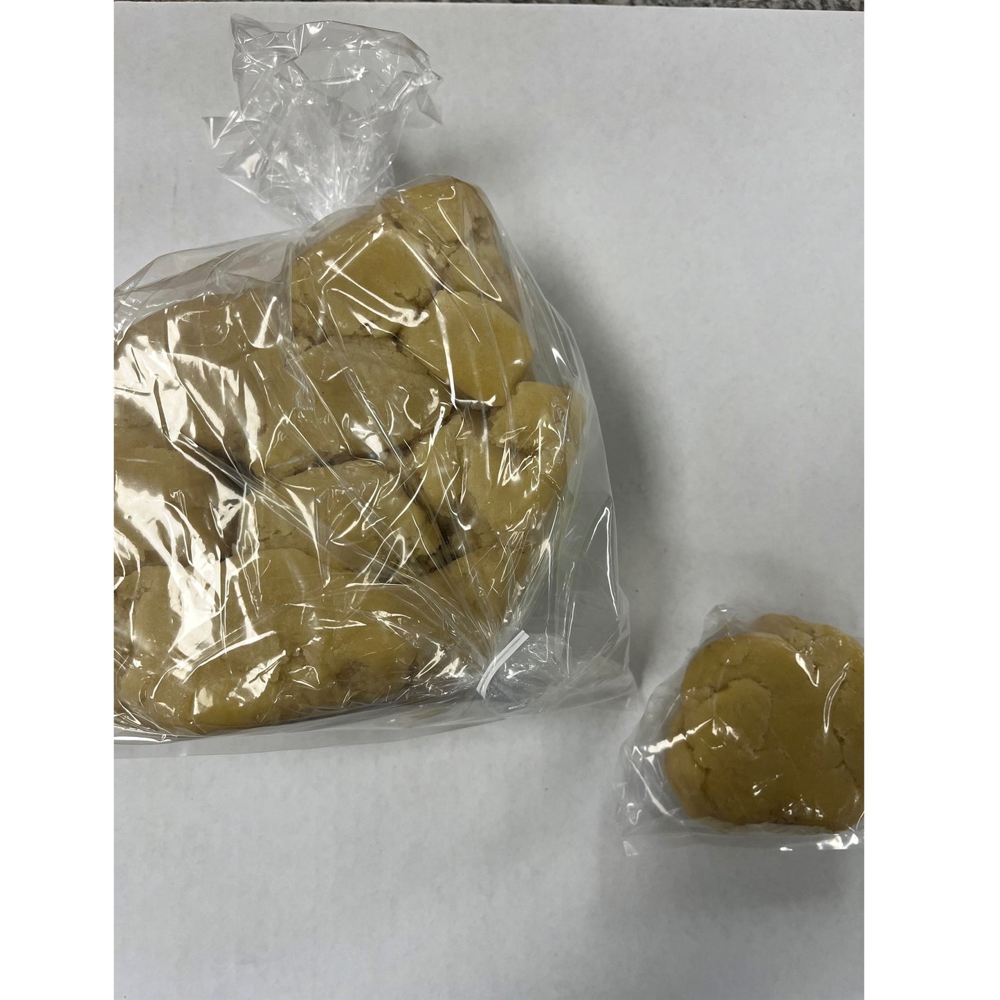 The image depicts a clear plastic bag containing a large quantity of almond paste, with a smaller portion of the paste packaged separately outside the main bag. The almond paste has a smooth, uniform texture and a pale golden color, characteristic of this confectionery ingredient often used in baking and pastry making.