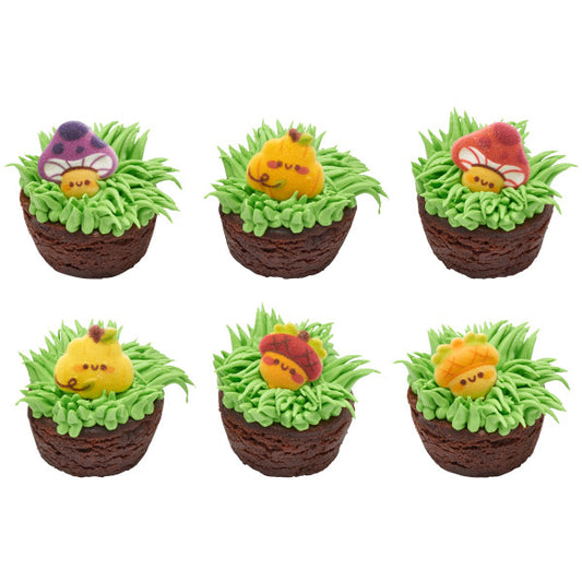 Chocolate brownie cupcakes topped with green grass-like frosting, each featuring a colorful edible sugar decoration of mushrooms, pumpkins, or acorns for a playful autumn-themed dessert.
