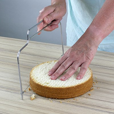 Person using PME cake leveller to cut even layers from a round sponge cake on a wooden surface.