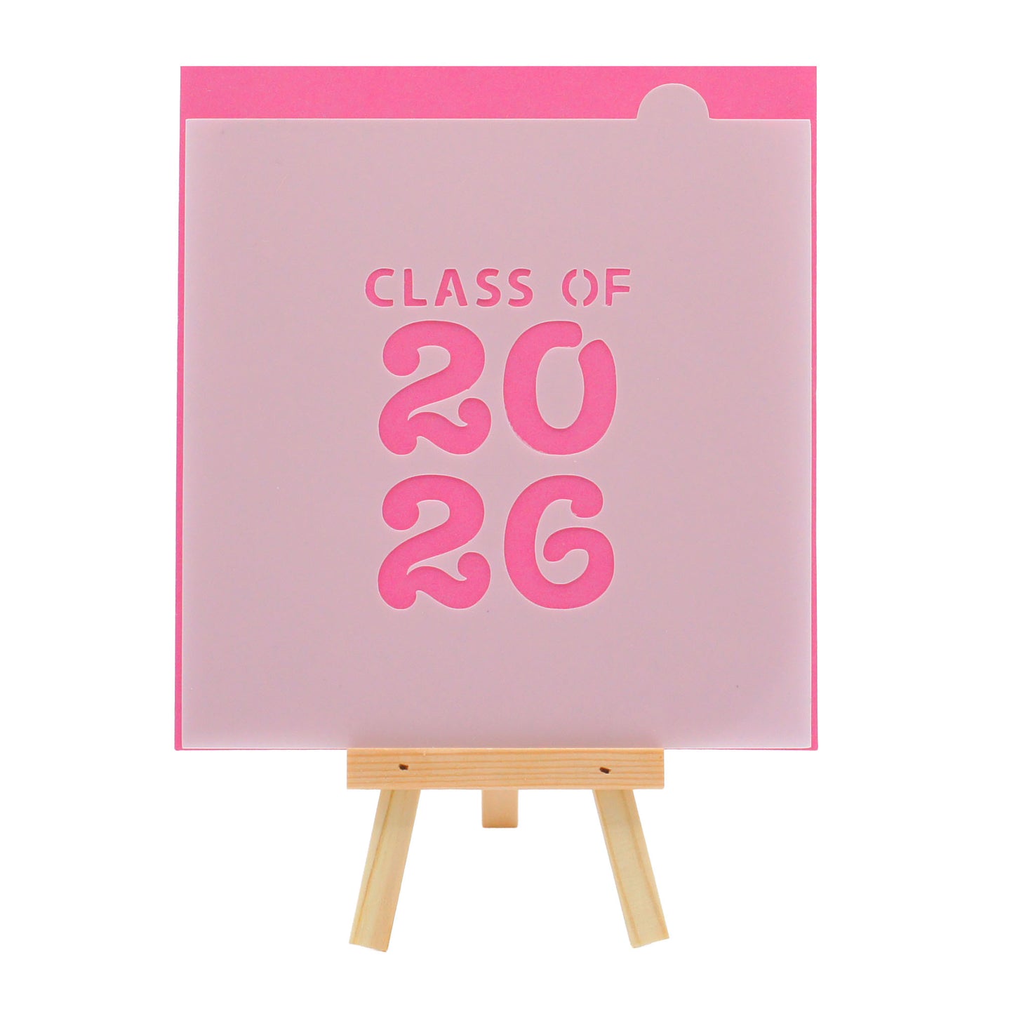 Translucent graduation cookie stencil with the words “CLASS OF 2026” in bold block text, displayed on a small wooden easel against a bright pink background.