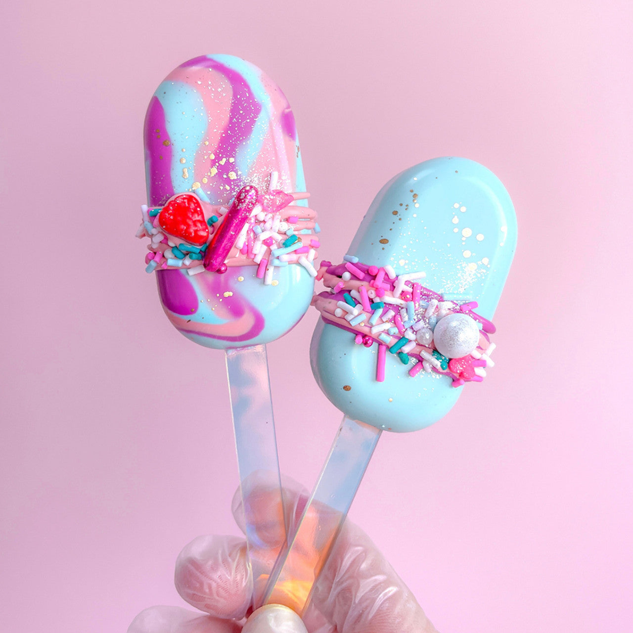 Pastel-dipped cakesicles coated in Colour Mill Baby Blue and swirled designs, decorated with bright sprinkles on a pink background