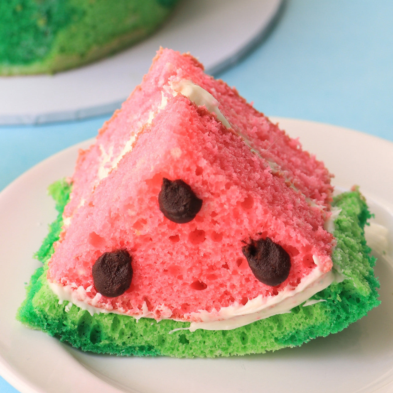 Slice of watermelon-themed cake featuring vibrant green and pink layers with chocolate chip “seeds.”