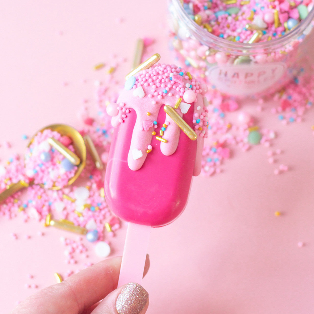 Bright pink cake pop decorated with pastel pink drip and colorful sprinkles, set against a pink background.