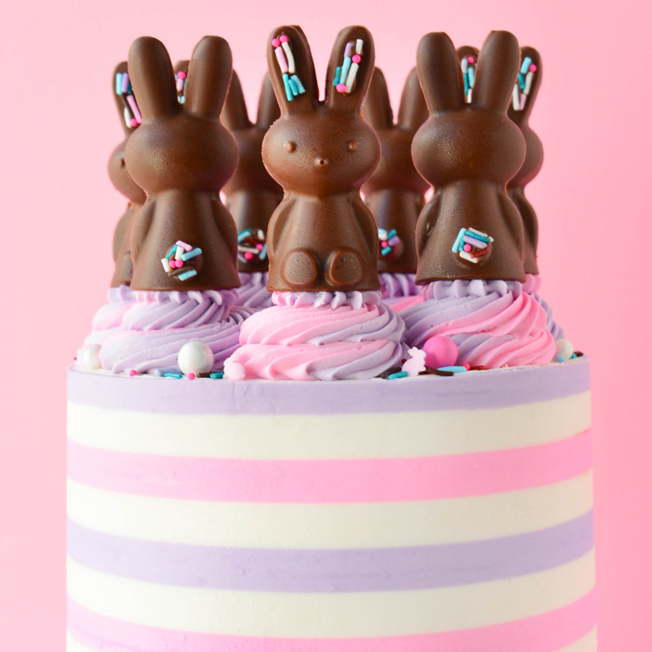 Striped cake in lavender, pink, and white topped with chocolate bunny toppers and swirled lavender buttercream.