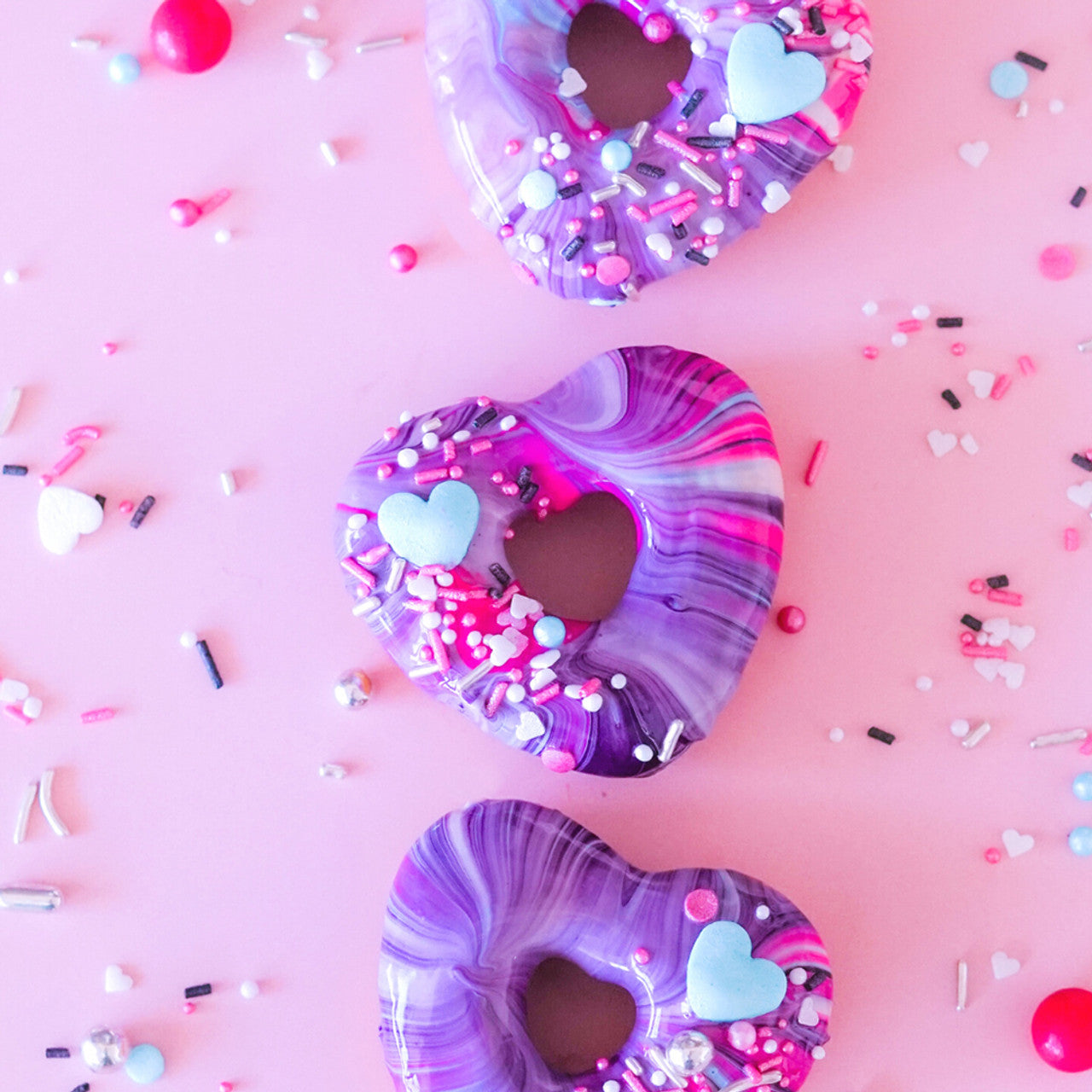 Heart-shaped donuts with vibrant purple and pink marbled glaze decorated with pastel blue heart candies and colorful sprinkles