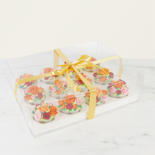 Assembled PME Crystal Cake Box holding 12 floral cupcakes, displayed in a clear box with gold satin ribbon on marble surface.