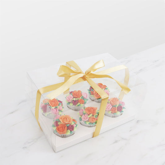 Clear PME Crystal Cake Box holding 6 floral cupcakes, tied with gold ribbon on a marble background.