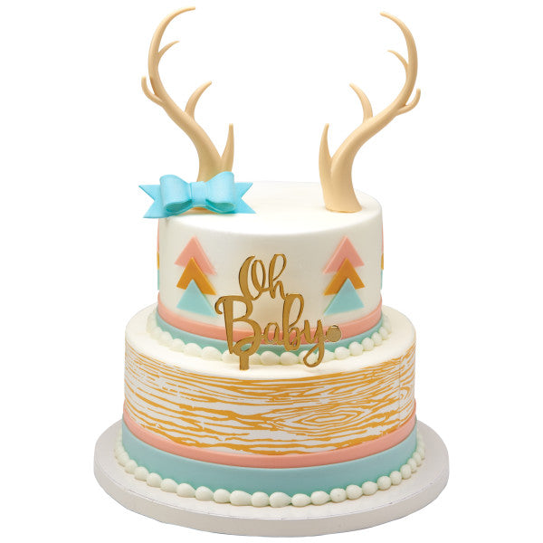 “Oh Baby” two-tier cake in pastel tones with a blue bow and deer antler toppers, perfect for baby showers.