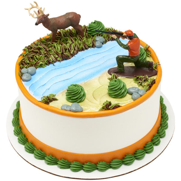 Round cake decorated with a forest and stream design, featuring a kneeling hunter figurine aiming at a standing buck topper.