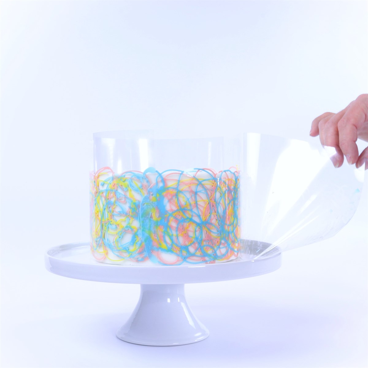 Clear acetate cake collar being peeled from a cake with colorful chocolate decoration on a white stand.