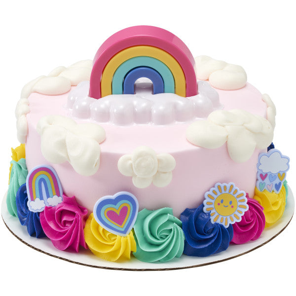 Round pink frosted cake with white cloud dollops and colorful buttercream rosettes around the base, topped with a stacked rainbow on a white cloud tray.