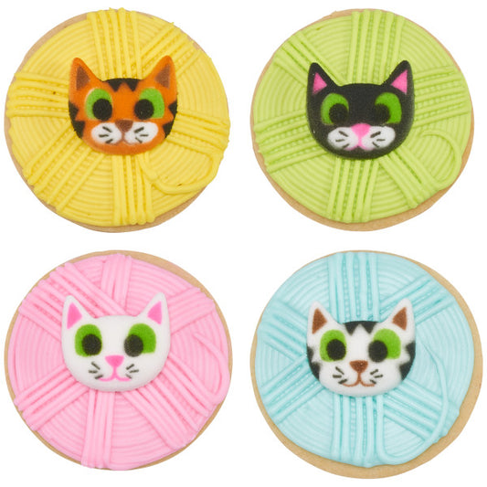 Four cookies decorated with yarn ball designs, each topped with a colorful kitten face edible decoration in orange, black, white, and mixed colors.