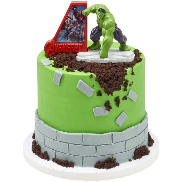 Round green cake styled like cracked concrete with gray brick banding and chocolate crumb rubble, topped with Hulk figure on base and a red Avengers sign.
