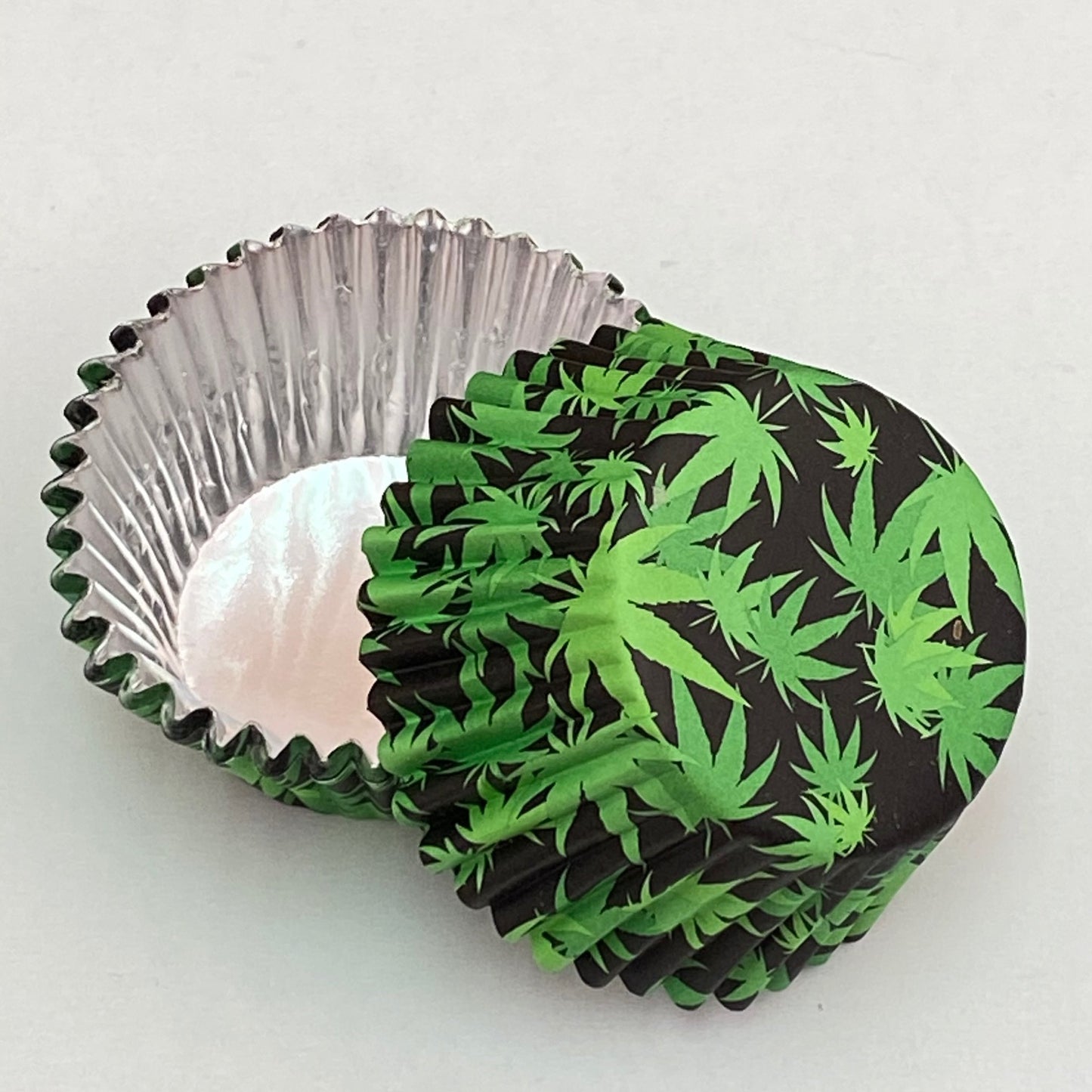 Foil cupcake liners with green cannabis leaf print on black paper exterior and shiny silver foil interior, shown stacked and separated for detail.