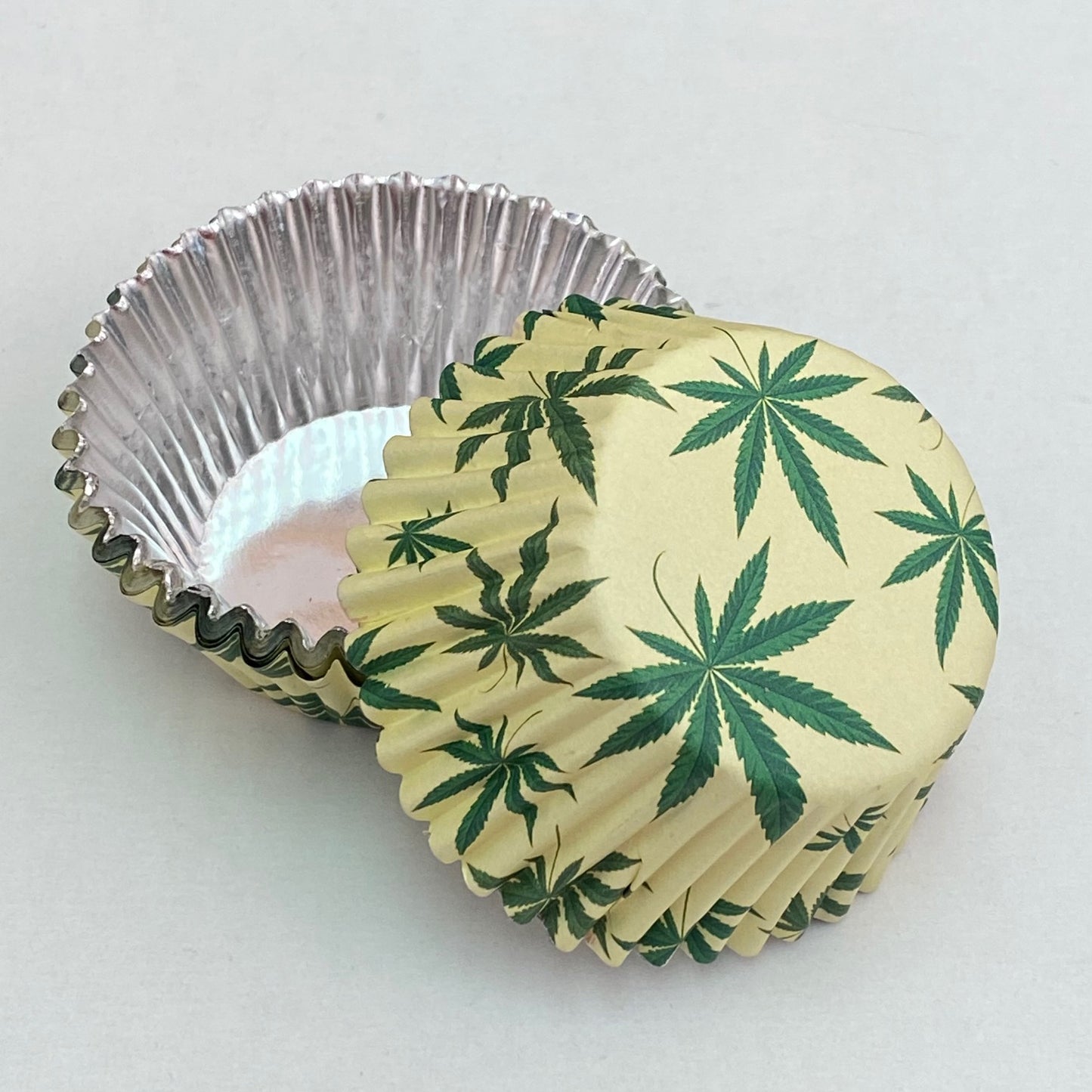 Foil cupcake liners with green cannabis leaf print on cream paper exterior and shiny silver foil interior, shown stacked and separated for detail.