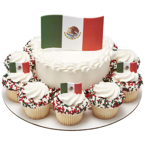 Round white cake with white frosting, surrounded by cupcakes topped with red, green, and white sprinkles and Mexican flags for a fiesta celebration.