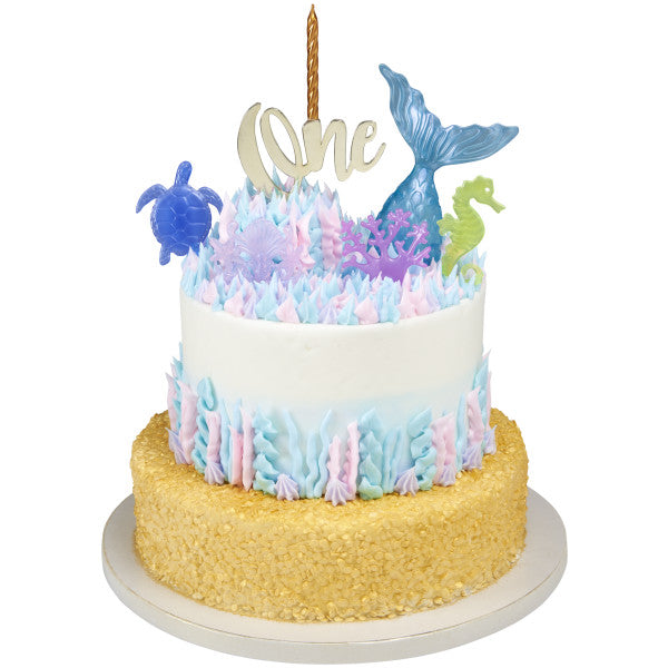 Two-tier cake with ocean-themed frosting and gold bottom tier, topped with Mystical Mermaid plastic toppers and a gold "One" candle decoration.