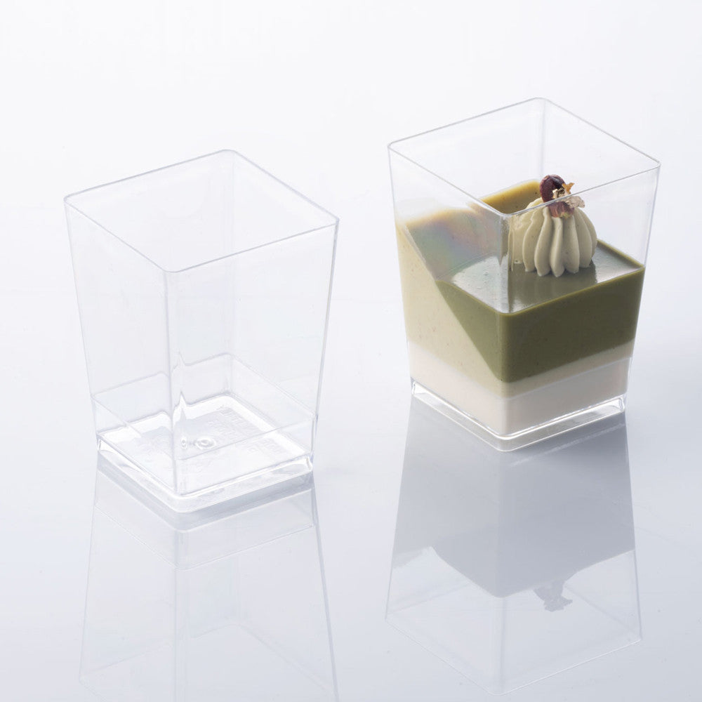 Two clear mini square plastic dessert cups, one empty and one filled with layered green and cream dessert topped with a piped dollop and garnish, on a white background with reflections.