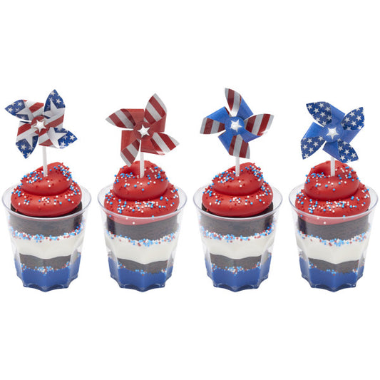 Four layered dessert cups with red frosting swirls and patriotic sprinkles, each topped with a red, white, and blue pinwheel cupcake pick.