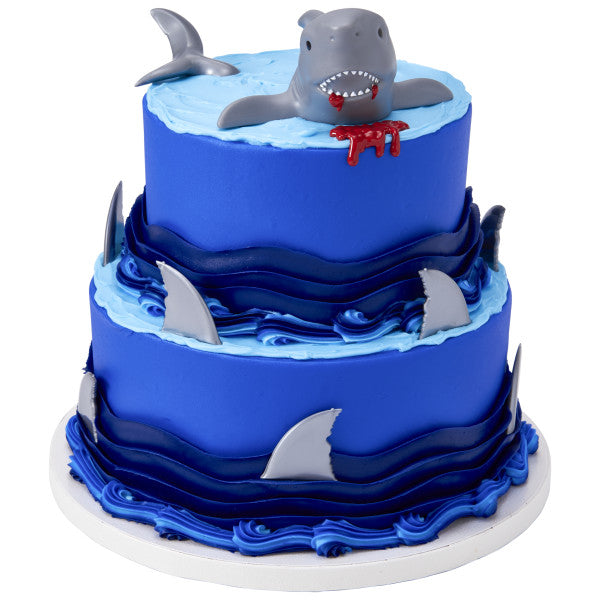 Tiered cake decorated with shark fin toppers, waves, and a 3D shark figure emerging from the top layer