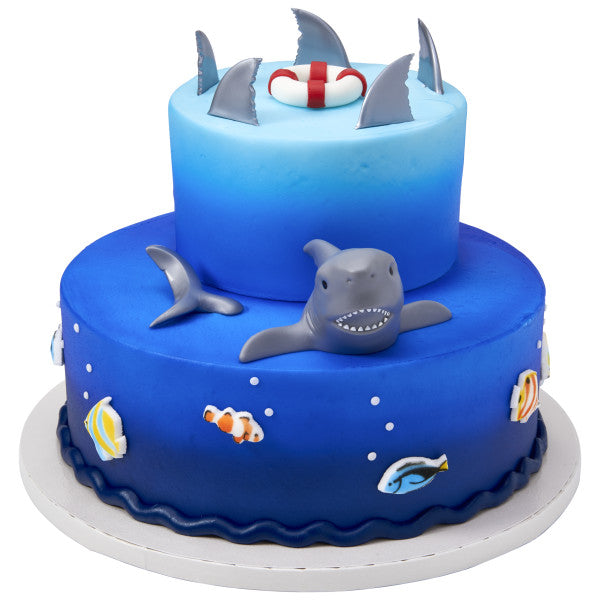 Ocean-themed cake featuring shark fin toppers, a shark figure, and colorful fish decorations for a beach or sea party