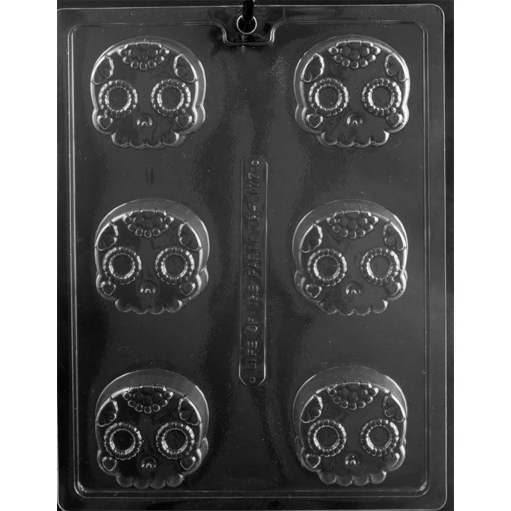 Clear plastic chocolate mold with six skull-shaped cavities, each featuring a decorative sugar skull face with raised circles around the eyes and dotted floral-style details on the forehead and cheeks.