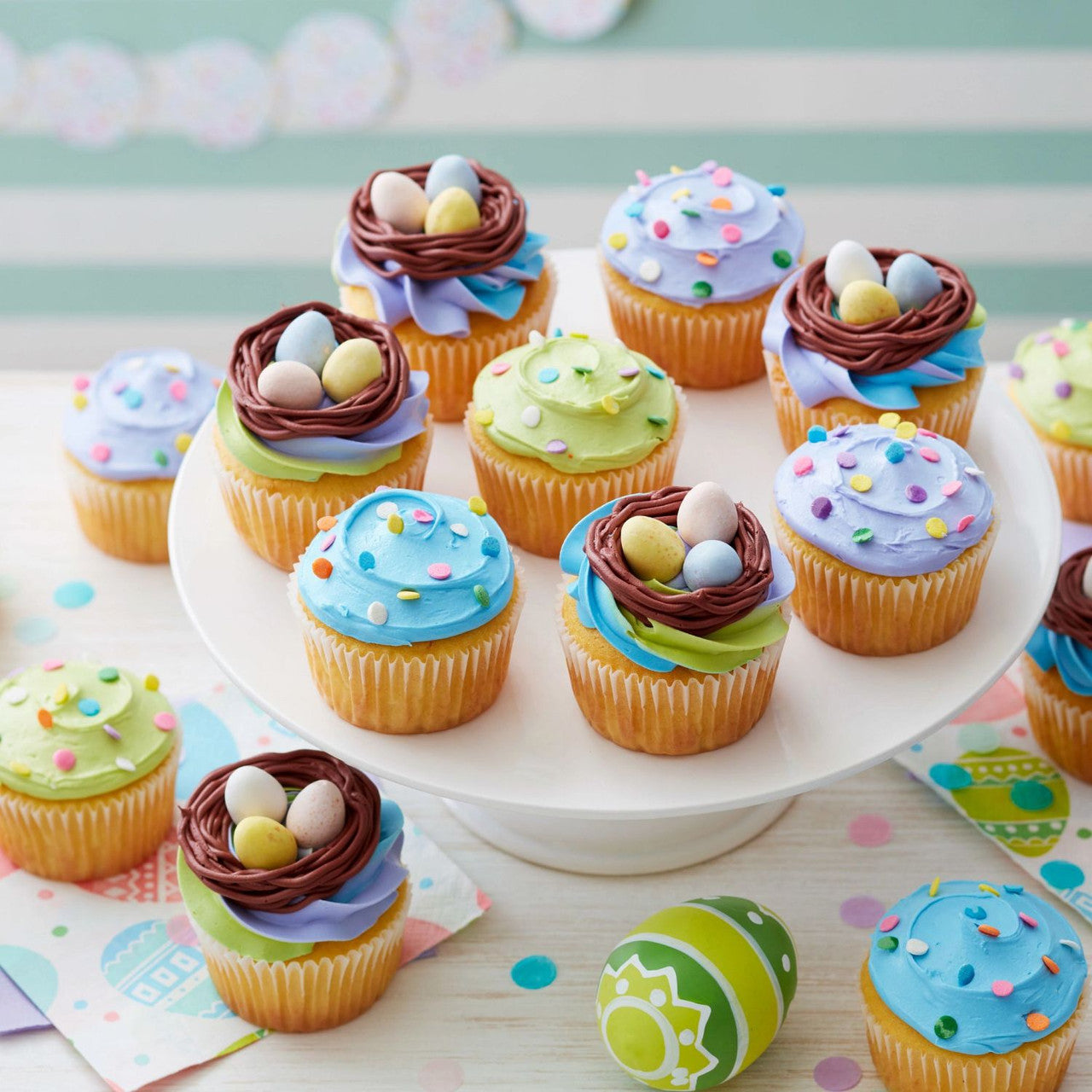 Pastel frosted cupcakes in colorful baking cups on a white cake stand, decorated for spring.