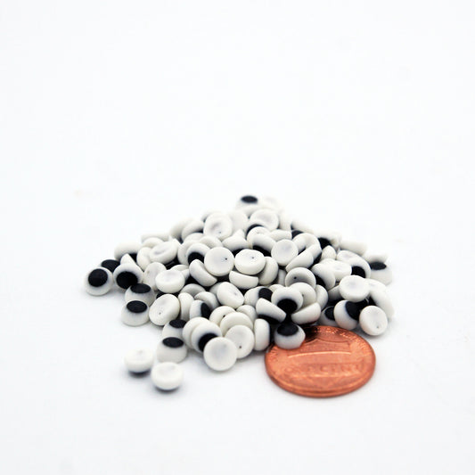 Pile of small white and black royal icing candy eyes next to a penny for scale, measuring approximately 3/16 inch, ideal for decorating cookies and cupcakes
