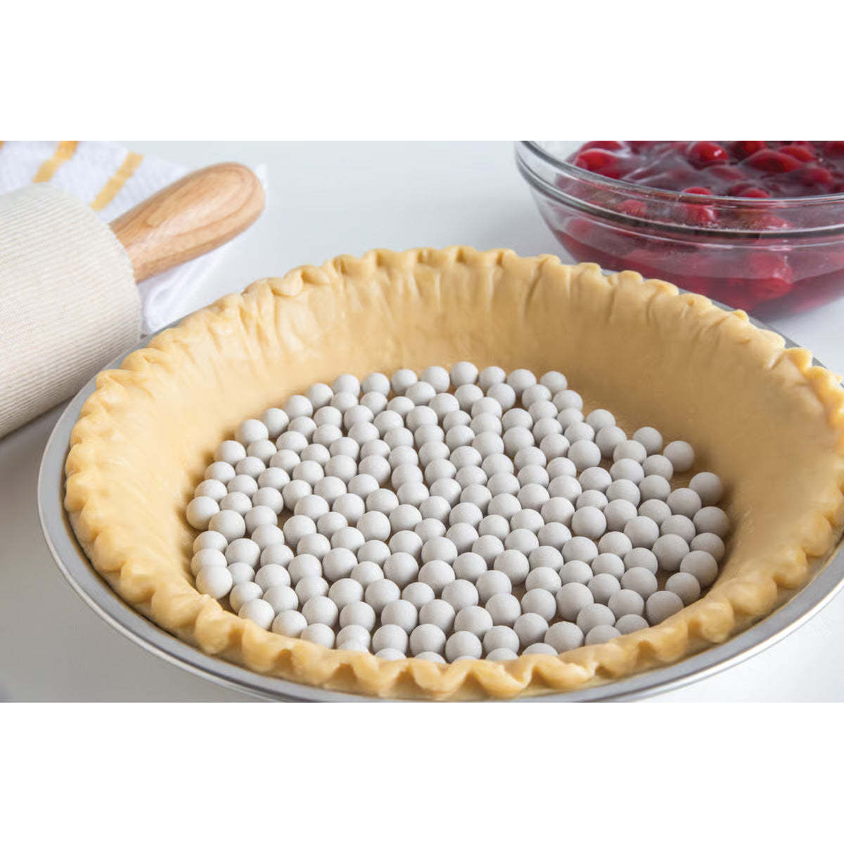 Unbaked pie crust filled with ceramic pie weights, ready for blind baking, with rolling pin and cherry filling in the background.