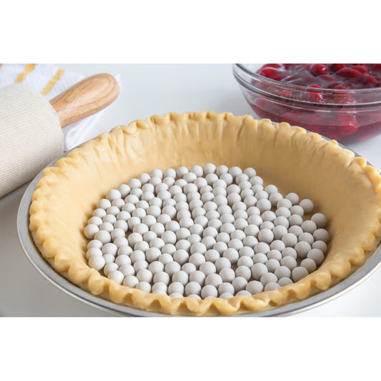Unbaked pie crust filled with ceramic pie weights, ready for blind baking, with rolling pin and cherry filling in the background.