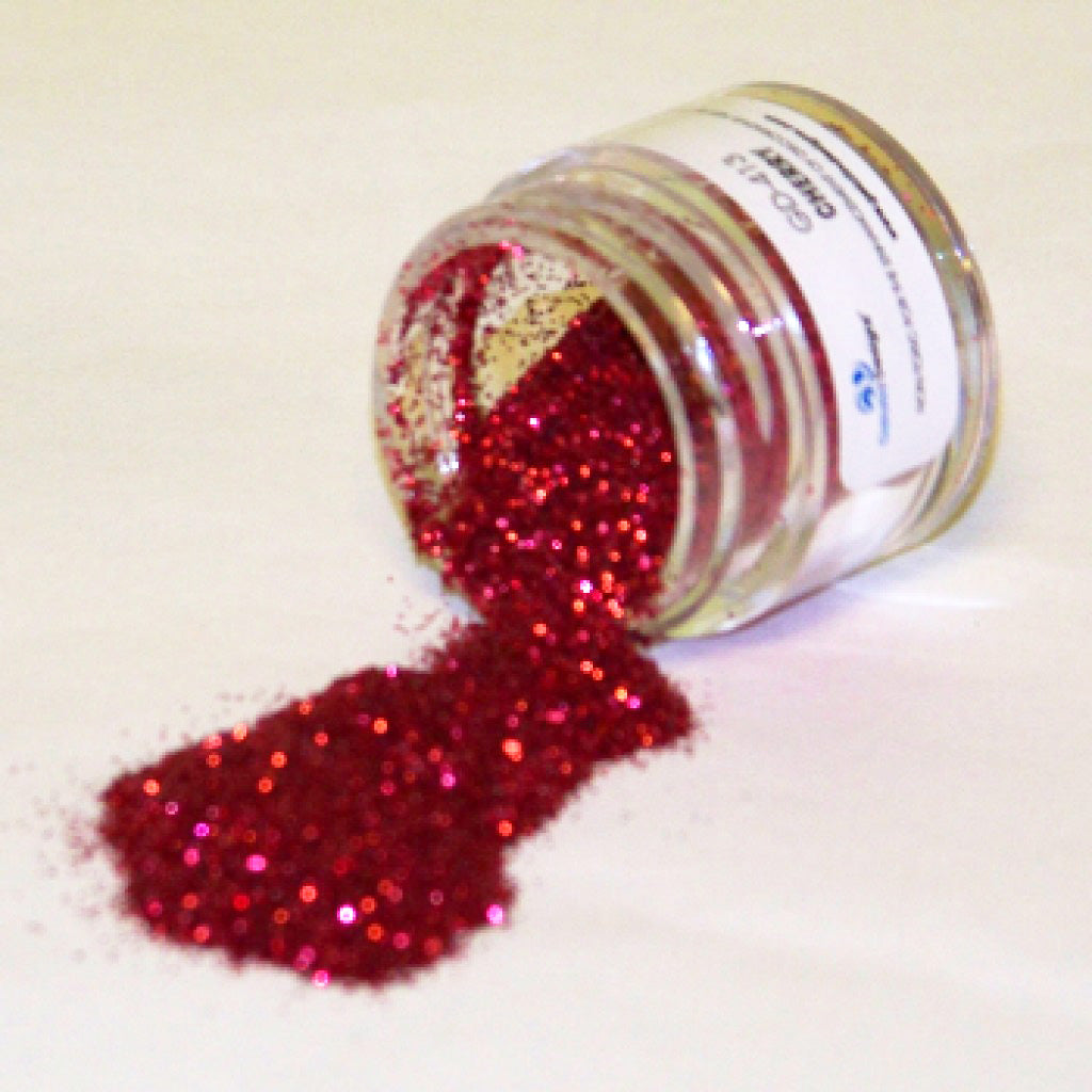 Small clear jar tipped on its side spilling bright cherry red glitter dust onto a white surface, with sparkling reflective particles visible throughout.