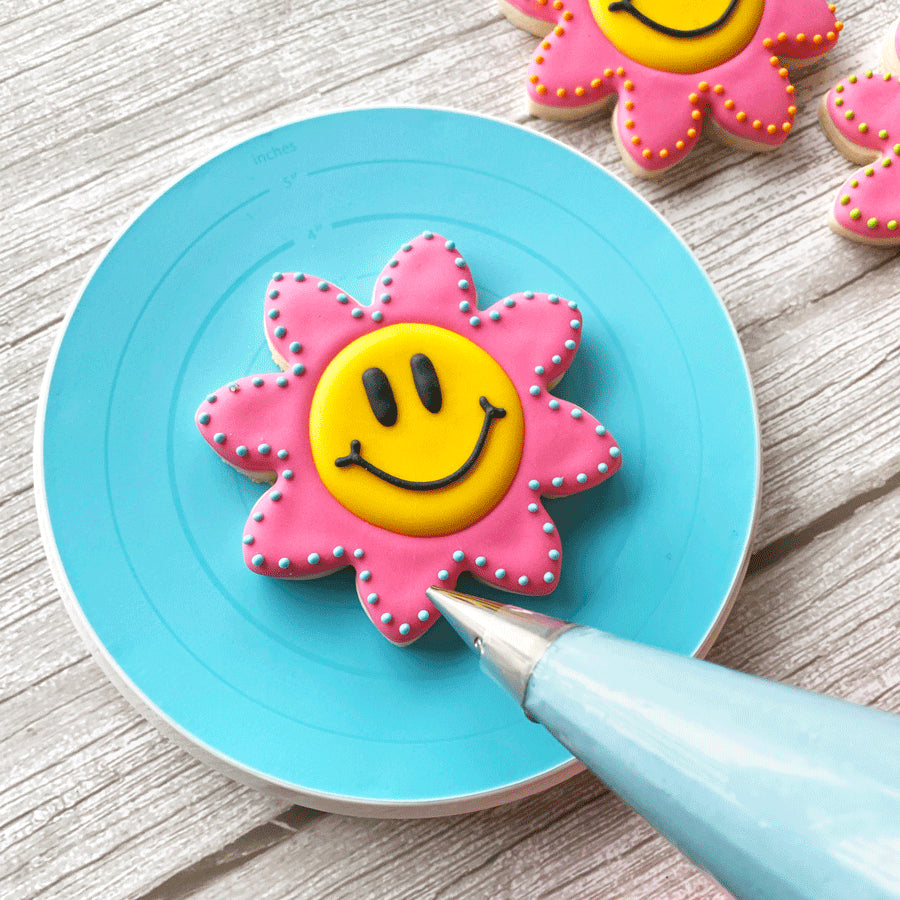 Smiley face flower cookie being decorated with blue icing on the Cookie Turntable.