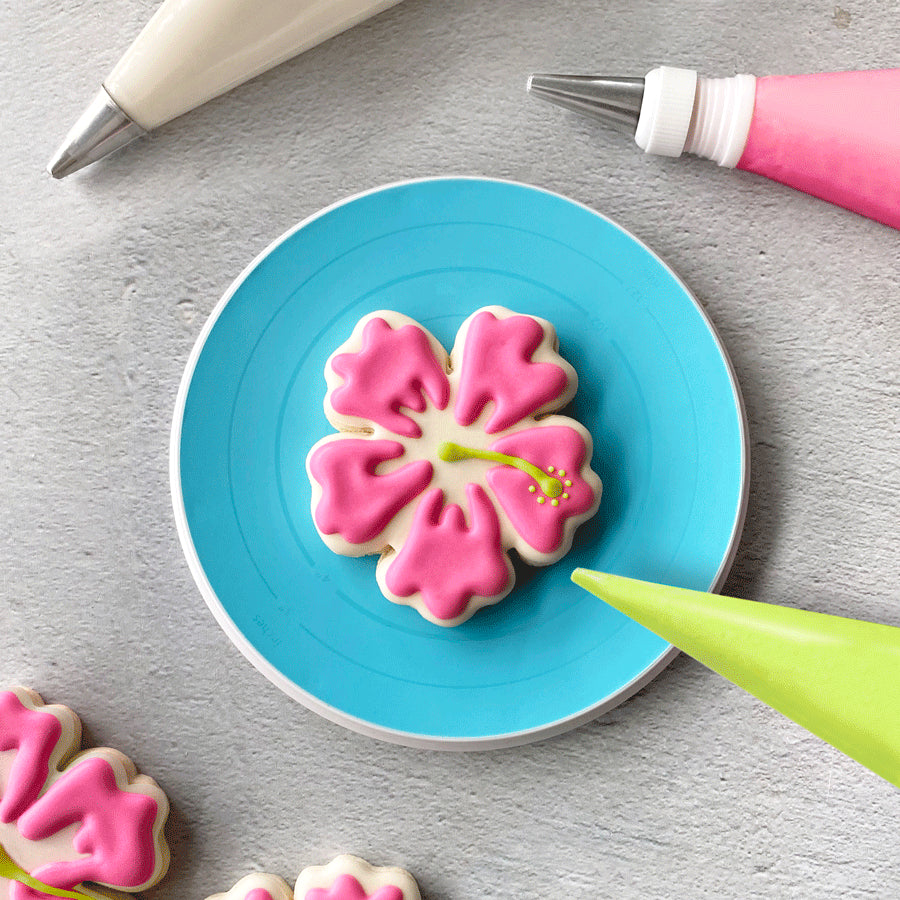 Cookie Turntable in use, holding a hibiscus-shaped cookie with pink and green icing.