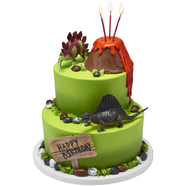 Two-tier green cake with a volcano and dinosaur toppers, including candles and a wooden-style “Happy Birthday” sign.