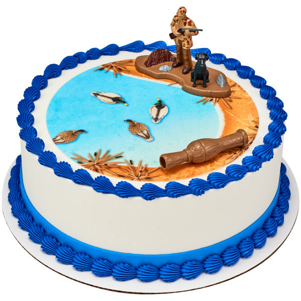 Round cake decorated with hunter and dog topper, duck call, and water-themed icing with duck prints.