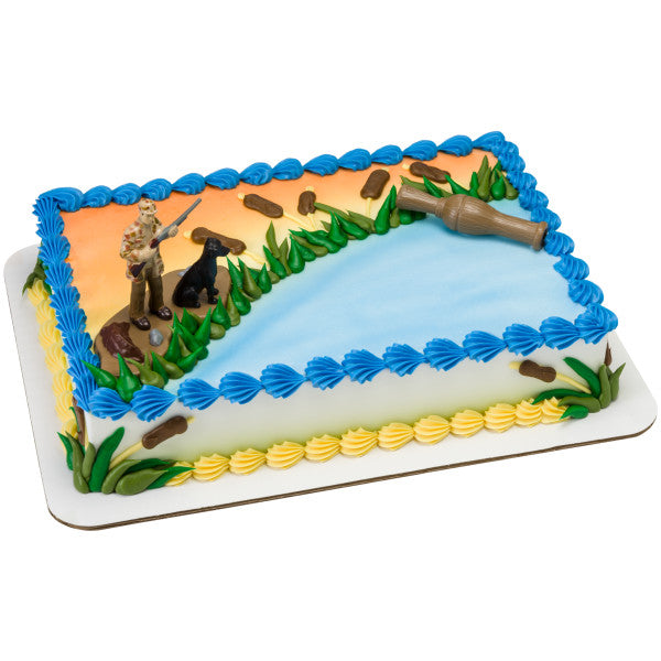 Side view of a rectangular cake decorated with hunting-themed figures and blue pond icing.