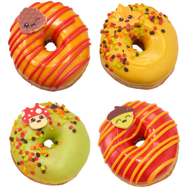 Four fall-themed donuts decorated with orange and green icing, sprinkles, and smiling pressed sugar toppers shaped like a mushroom, leaf, acorn, and pine cone.