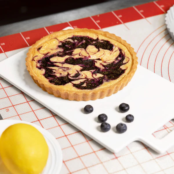 Baked tart with blueberry swirl filling in tart pan on a cutting board, surrounded by baking tools and fresh blueberries.
