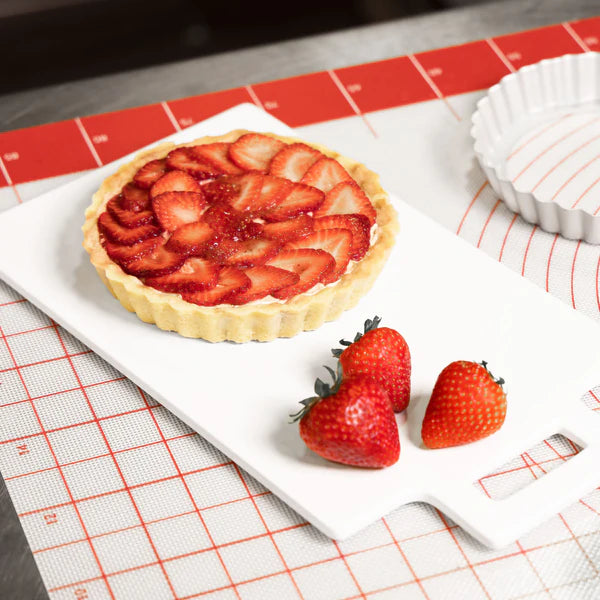 Strawberry tart in tart pan on a cutting board, with fresh strawberries placed alongside on a red and white silicone mat.