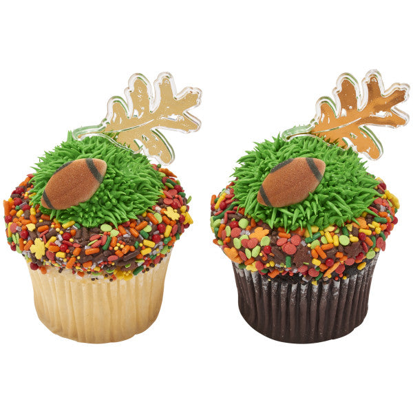 Two cupcakes decorated with green piped frosting to resemble grass, each topped with a brown sugar football and gold foil oak leaf pick, surrounded by colorful fall sprinkles.