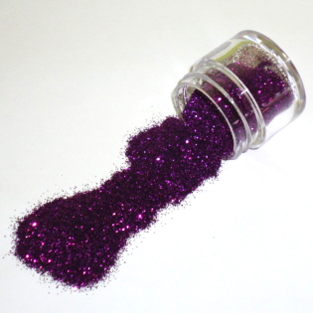 Small clear jar tipped on its side spilling grape purple glitter dust onto a white surface, with fine sparkling particles visible throughout.