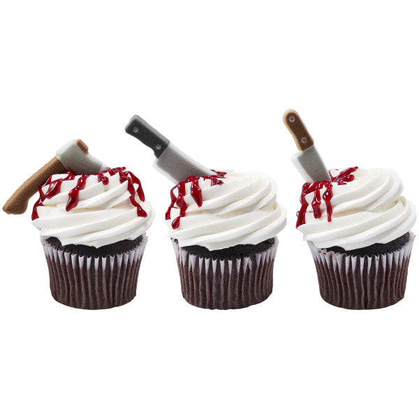 Three cupcakes topped with edible sugar axes, cleavers, and knives, with red icing resembling dripping blood on the white frosting.