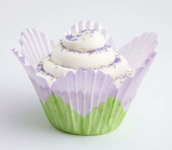 Lavender and green petal-shaped baking cup holding a frosted cupcake with white swirled icing and small purple sprinkles on a light background.