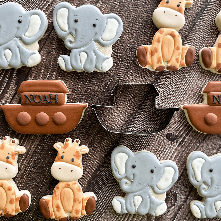 Decorated ark-shaped cookies with brown icing, designed to resemble Noah’s Ark, displayed on a wooden surface alongside decorated elephant and giraffe cookies.