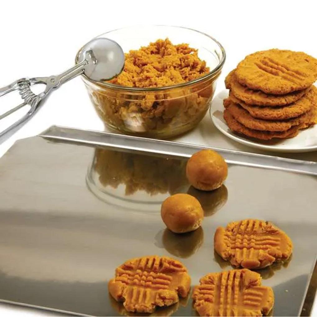 Number 24 stainless steel cookie scoop beside a bowl of dough, rounded portions, and fork-marked cookies on a metal cookie sheet.